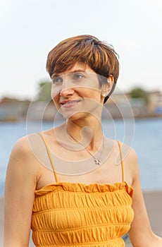 Portrait of cheerful middle-aged lady standing by river in city embankment, enjoing summer