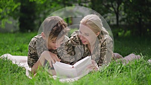 Portrait of cheerful military mother reading book with teenage son lying on sunny meadow in summer spring park. Happy