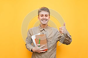 Portrait of a cheerful nerd in a shirt and glasses stands on a yellow background with books and exercise books in his hands, looks