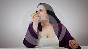 Portrait of cheerful obese Caucasian woman sitting at the table and eating apple. Smiling brunette girl eats healthy low