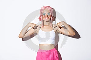 Portrait of cheerful party girl with pink wig and bright makeup, pointing at herself and smiling confident, showing her