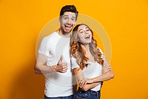 Portrait of cheerful people man and woman in basic clothing smiling and showing thumb up at camera, isolated over yellow