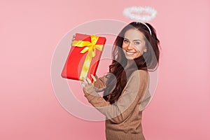 Portrait of cheerful pretty angelic young woman with halo holding big present and smiling to camera, satisfied with gift