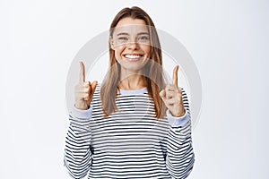 Portrait of cheerful pretty female model, smiling white teeth, showing direction to advertisement, pointing fingers up