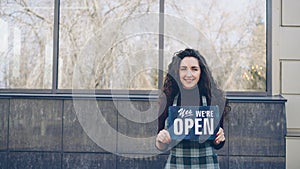 Portrait of cheerful pretty waitress in apron holding Yes We Are Open sign standing outside and smiling looking at