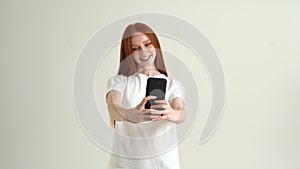 Portrait of cheerful pretty young woman taking selfie and smiling using smartphone standing on white isolated background
