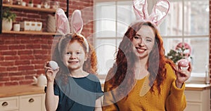 Portrait of cheerful redhead little kid girl with her mom playing with easter egg on the kitchen background. They are