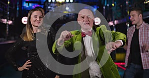 Portrait of cheerful senior Caucasian man and young pretty woman dancing in night club and looking at camera. Joyful old