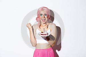 Portrait of cheerful smiling girl celebrating her birthday, wearing pink wig, holding b-day cake and shouting of joy