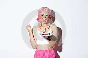 Portrait of cheerful smiling girl celebrating her birthday, wearing pink wig, holding b-day cake and shouting of joy