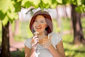 Smiling woman standing under trees in park or forest with big ice-cream and showing thumb up. Enjoying moment and summer leisure