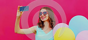 Portrait cheerful smiling woman taking selfie picture by smartphone with colorful balloons on a pink background