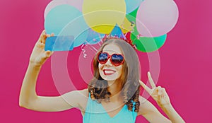 Portrait of cheerful smiling woman taking selfie picture by smartphone with colorful balloons on pink background