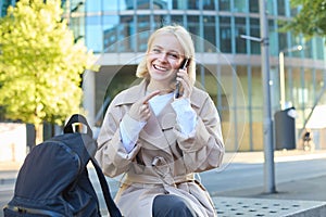 Portrait of cheerful smiling woman, talking on mobile phone, pointing at her smartphone while calling someone, smiling