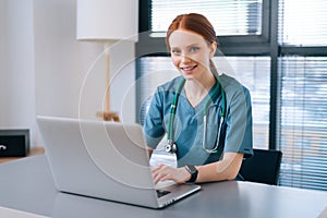 Portrait of cheerful smiling young female doctor in blue green medical uniform typing on laptop computer