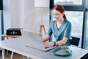 Portrait of cheerful smiling young female doctor in blue green medical uniform typing on laptop computer looking on