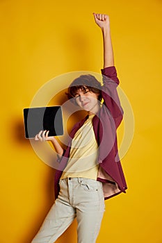 Portrait of cheerful teen girl with curly brown short hair posing with tablet isolated over yellow background. Online