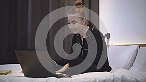 Portrait of cheerful teenage girl using video chat on laptop in bedroom. Happy relaxed Caucasian teenager smiling and