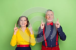 Portrait of cheerful two pensioners look indicate fingers up empty space isolated on green color background