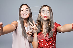 Portrait of cheerful two women friends make selfie by phone showing peace gesture standing isolated over white background