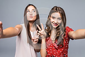 Portrait of cheerful two women friends make selfie by phone showing peace gesture standing isolated over white background