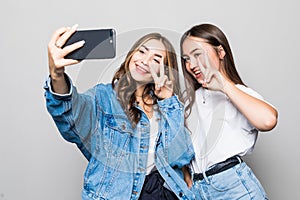 Portrait of cheerful two women make selfie by phone showing peace gesture. friends standing isolated over gray background