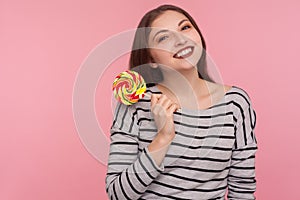 Portrait of cheerful woman in striped sweatshirt holding sweet round rainbow candy lollipop, pink background