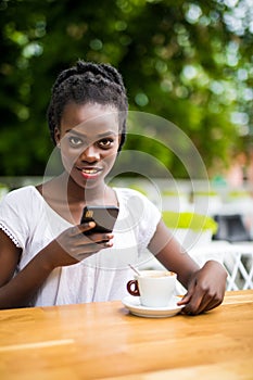 Portrait of cheerful young african woman sitting at outdoor cafe and tyoing on phone