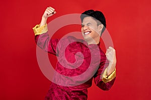Portrait of a cheerful young Asian man in red cheongsam qipao. He raised his fist as a gesture of celebration on a red background