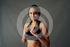 Portrait of cheerful young biracial woman in sports bra holding jump rope over shoulders in studio