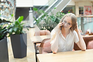 Portrait of a cheerful young caucasian blond business woman sitting on the table, talking at phone, smiling and looking away.