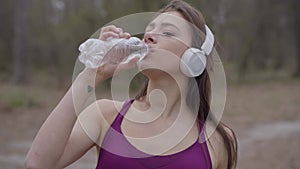 Portrait of cheerful young Caucasian sportswoman in headphones drinking water during workout break outdoors. Smiling