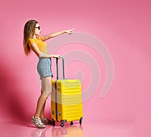 Portrait of a cheerful young caucasian woman dressed in summer clothes, with a suitcase and pointing finger away