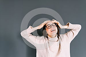 Portrait of cheerful young caucasian woman in eyeglasses holding hands on head on gray background looking on camera