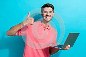 Portrait of cheerful young guy in pink t shirt shows thumb up like symbol holding laptop xiaomi isolated on aquamarine