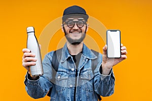 Portrait of cheerful young hipster, holding steel thermo eco bottle for water and smartphone with white mockup, on yellow.