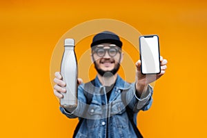 Portrait of cheerful young hipster, holding steel thermo eco bottle for water and smartphone with white mockup, on yellow.