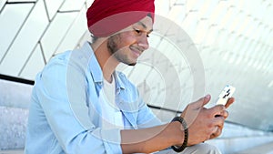 Portrait of cheerful young Indian guy in turban sending message on mobile phone