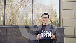 Portrait of cheerful young man in apron catering business owner holding `yes we are open` sign standing outside opposite