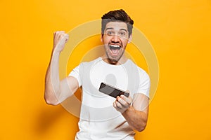 Portrait of a cheerful young man playing games on mobile phone isolated over yellow background