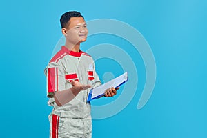 Portrait of a cheerful young mechanic holding clipboard and pointing with palm at camera isolated on blue background