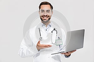 Portrait of cheerful young medical doctor with stethoscope working in clinic and holding laptop