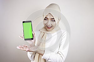 Portrait of cheerful young Muslim Asian woman pointing to cellphone with green screen