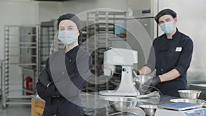 Portrait of cheerful young slim woman in Covid-19 face mask and cook uniform looking at camera smiling with blurred man