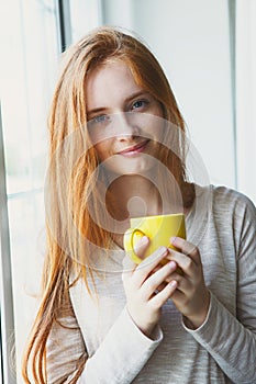 Portrait of cheerful young smiling red hair woman holding yellow coffee cup, enjoying morning coffee and new day, waking up