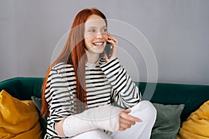 Portrait of cheerful young woman with broken injured arm wrapped in white gypsum bandage talking on smartphone with