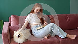Portrait of cheerful young woman reading book sitting on couch, stroking pretty miniature spitz dog in living room with