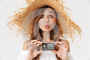 Portrait of cheerful young woman 20s wearing big straw hat rejoicing while holding credit card, isolated over white background