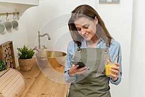 Portrait of cheerful young woman standing in kitchen with cup of fresh orange juice and checking text messages on her