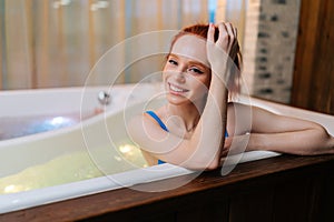 Portrait of cheerful young woman in swimsuit relaxed sitting in bath with hydromassage at luxury spa salon, looking at